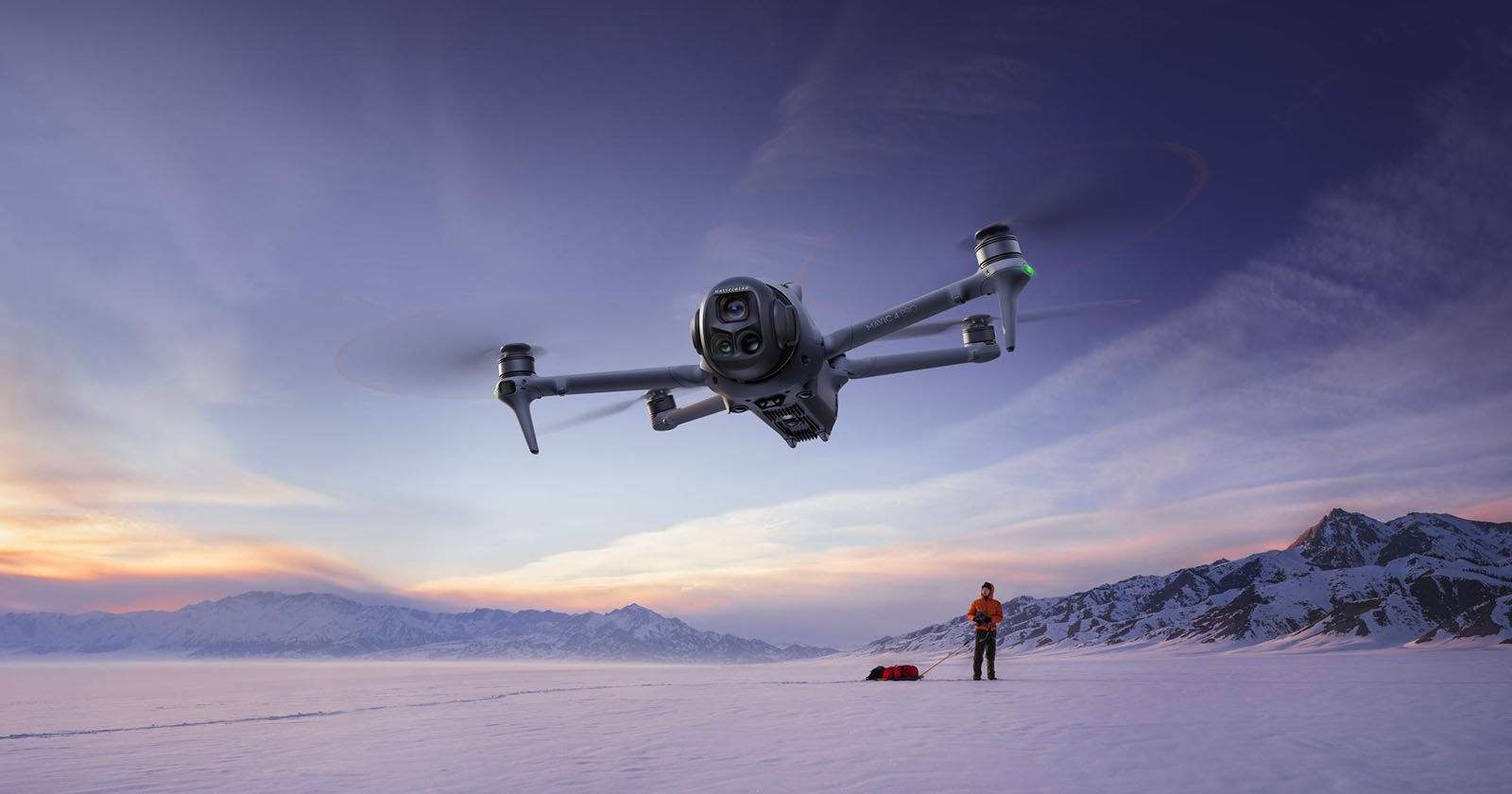 A large drone flies close to the camera over a snowy landscape at sunset; in the background, a person stands near a sled with mountains visible in the distance.