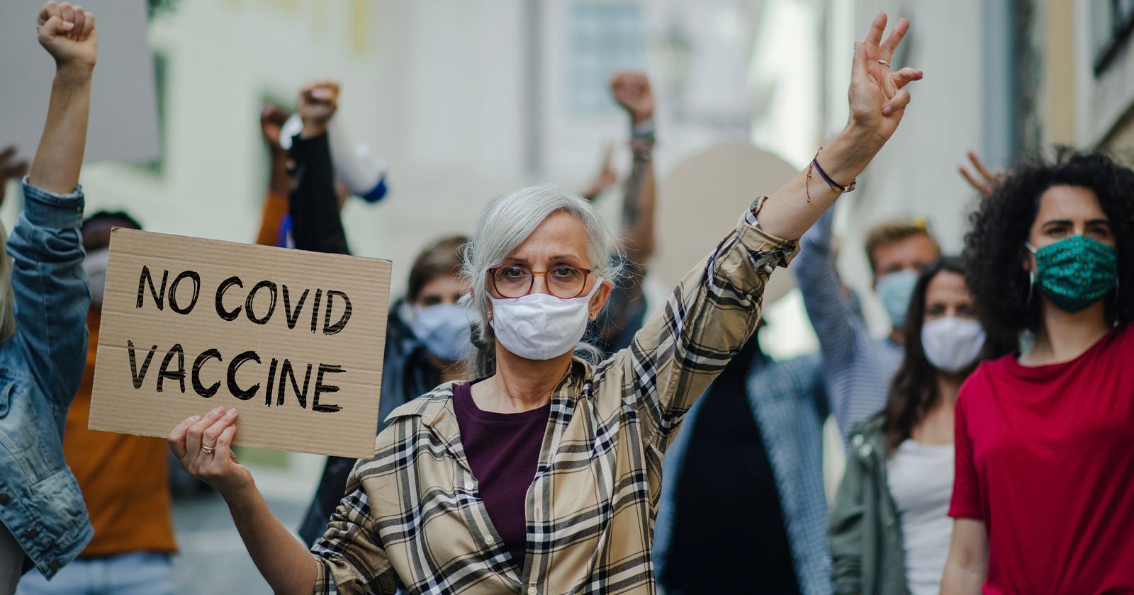 A group of people are protesting outdoors. A woman in the foreground with glasses and a mask holds up a sign reading NO COVID VACCINE. Others in the background also have raised fists and are wearing masks.