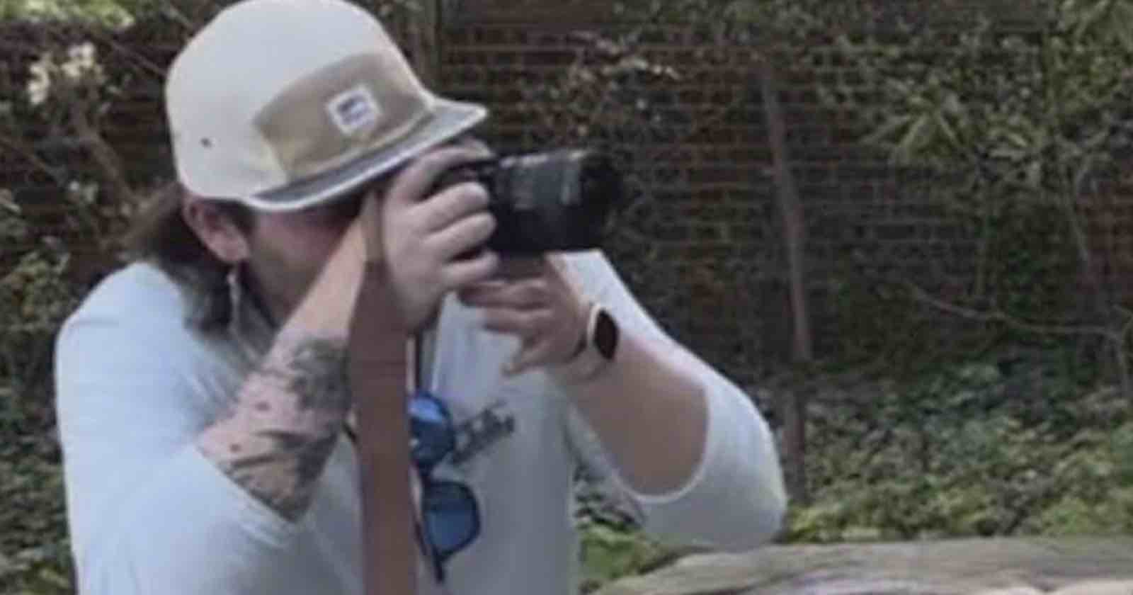 A person wearing a beige cap and white shirt is holding a camera up to their face, taking a photo outdoors with greenery and a brick wall in the background.