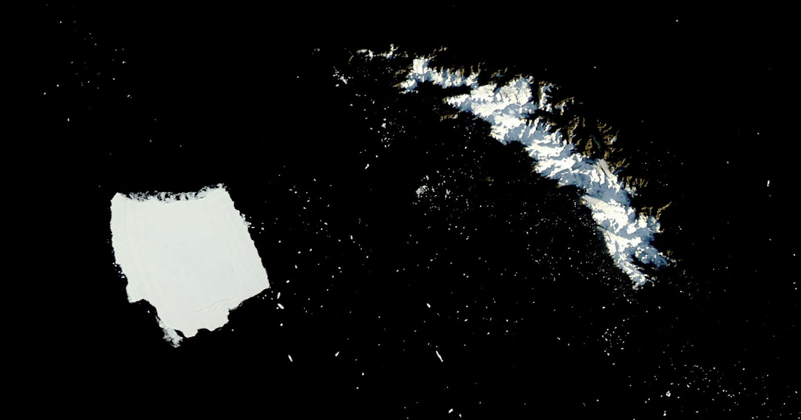 A large rectangular iceberg floats near a rugged, snow-covered island in a dark expanse of ocean, viewed from above.