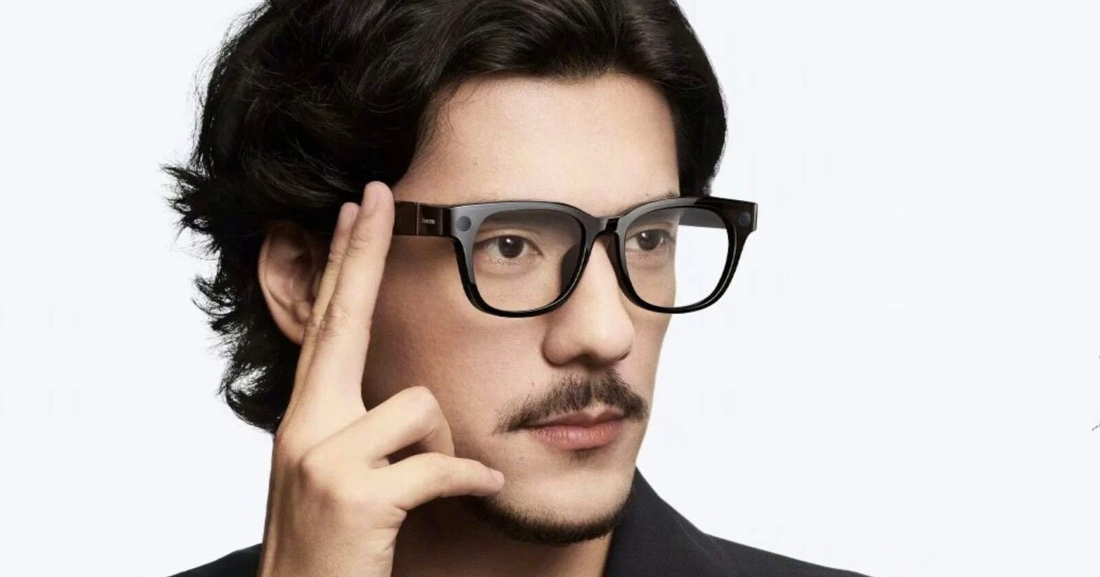 A man with black hair and a mustache is wearing smart glasses and touching the side of the frame with his fingers, as if using a touch control. The background is plain and light-colored.