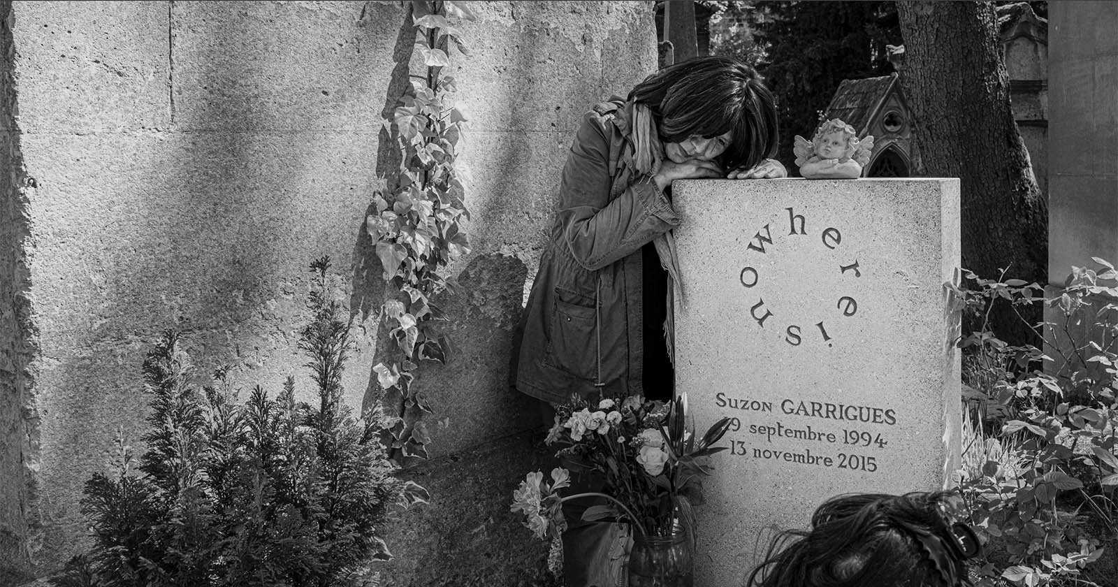 A woman leans on a gravestone, appearing to grieve. The gravestone reads Nowhere! Us! and marks the grave of Suzon Garrigues (1994–2015). Flowers and a small angel statue are placed in front of the stone.
