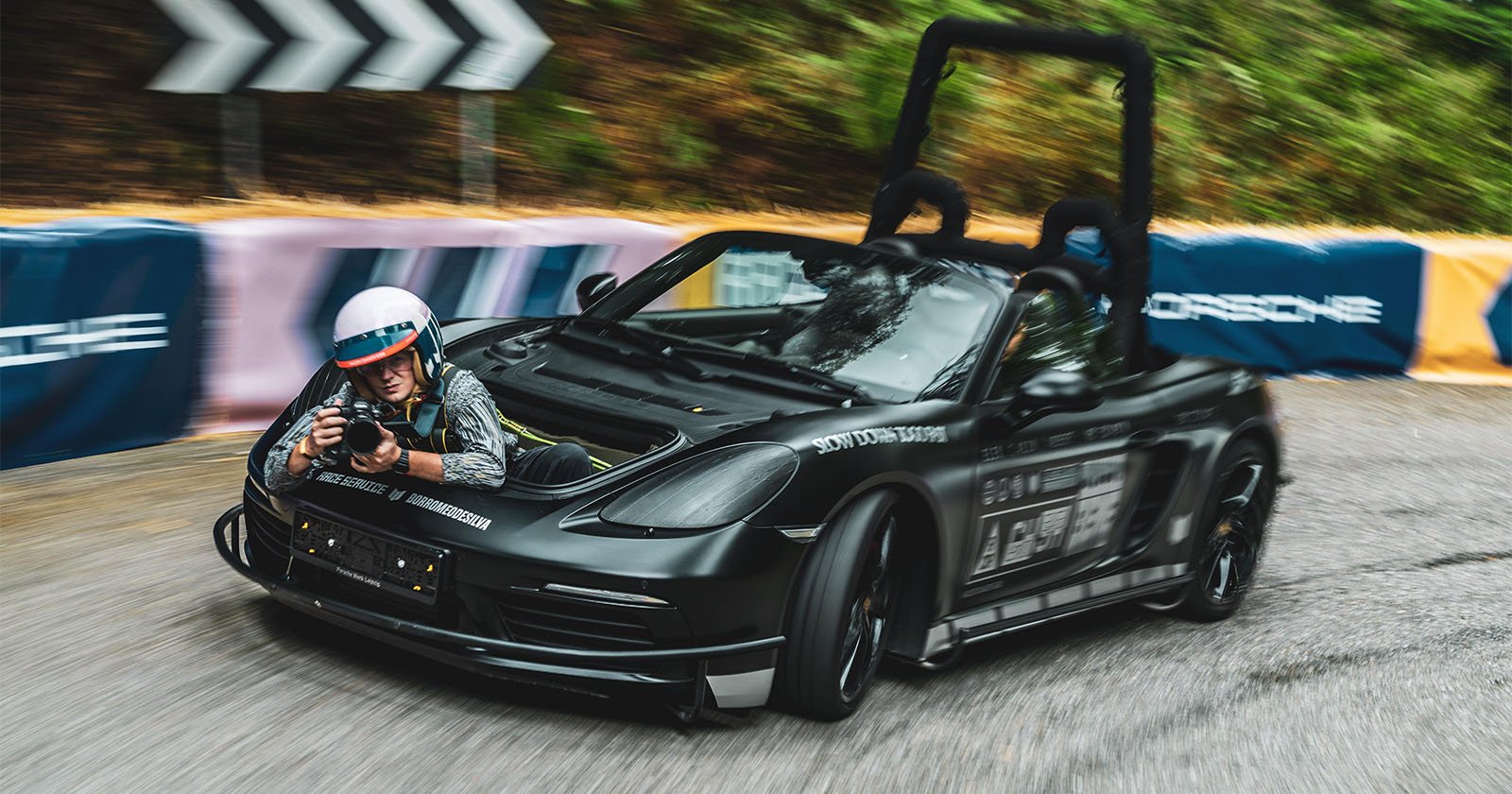 A black convertible car with a mounted camera rig and a person in a helmet lying where the hood should be, holding a camera, speeds around a sharp curve on a racetrack bordered by colorful barriers.