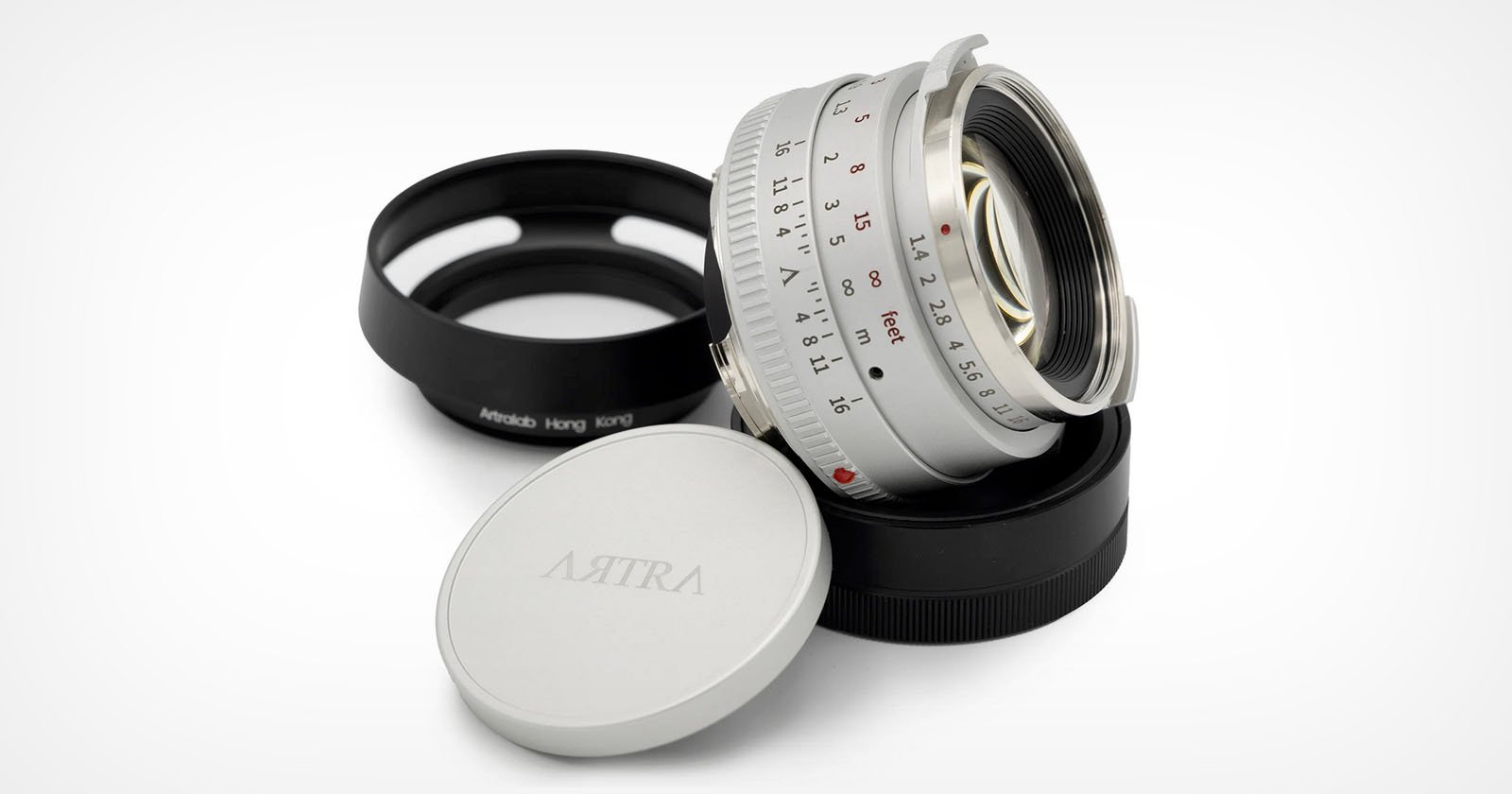 A silver camera lens with a visible focus and aperture ring, resting on a black lens cap labeled Artra and another cap in the background. The lens displays aperture markings from f/1.1 to f/16.
