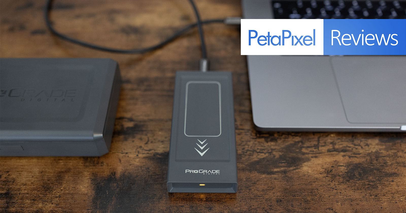 An external storage device connected to a laptop via a USB-C cable is shown on a wooden surface. The image also includes a ProGrade Digital card holder on the left side. The top-right text overlay reads PetaPixel Reviews.