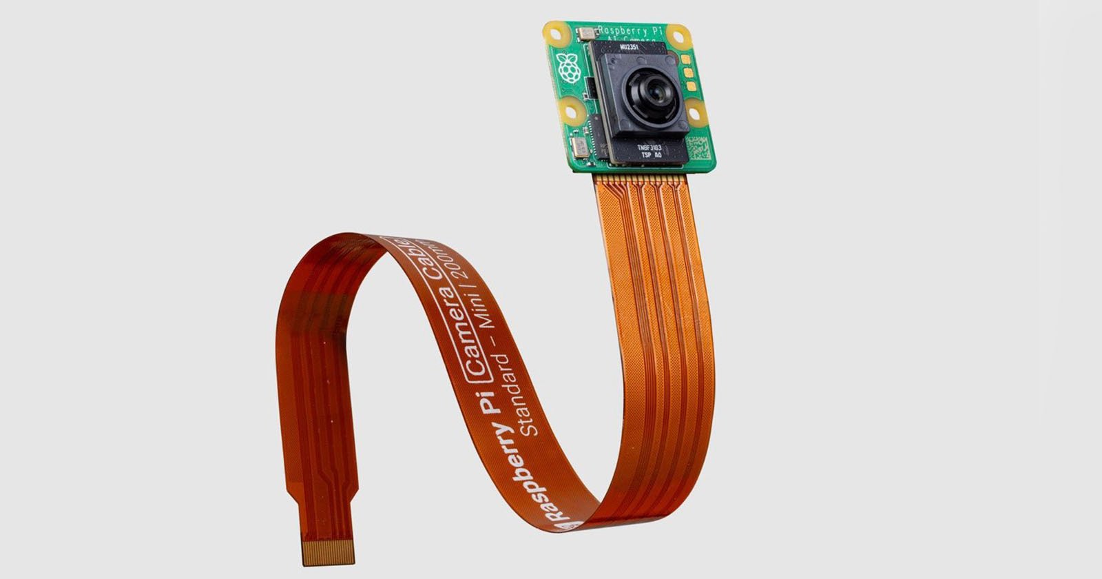 A Raspberry Pi camera module attached to a flexible orange ribbon cable. The cable is extended and has text printed on it, with the camera lens and green circuit board visible at the end. The background is plain and light-colored.
