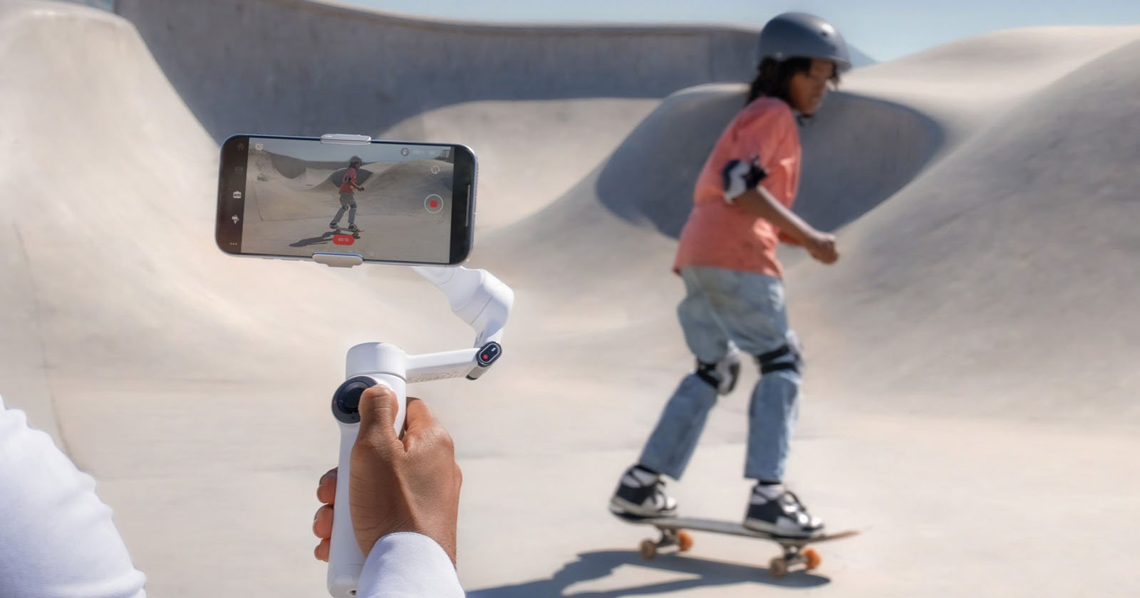 A person holds a smartphone mounted on a gimbal, recording a child wearing a helmet and pads skateboarding in a concrete skatepark.