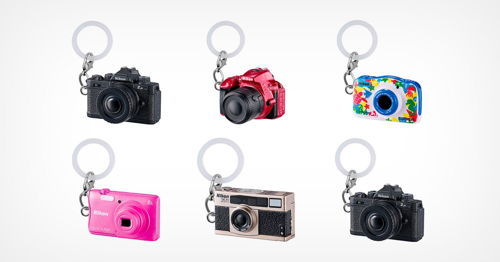 Six miniature camera-shaped keychains in different styles and colors, including black, red, blue with a colorful pattern, pink, silver, and black, arranged in two rows on a white background.