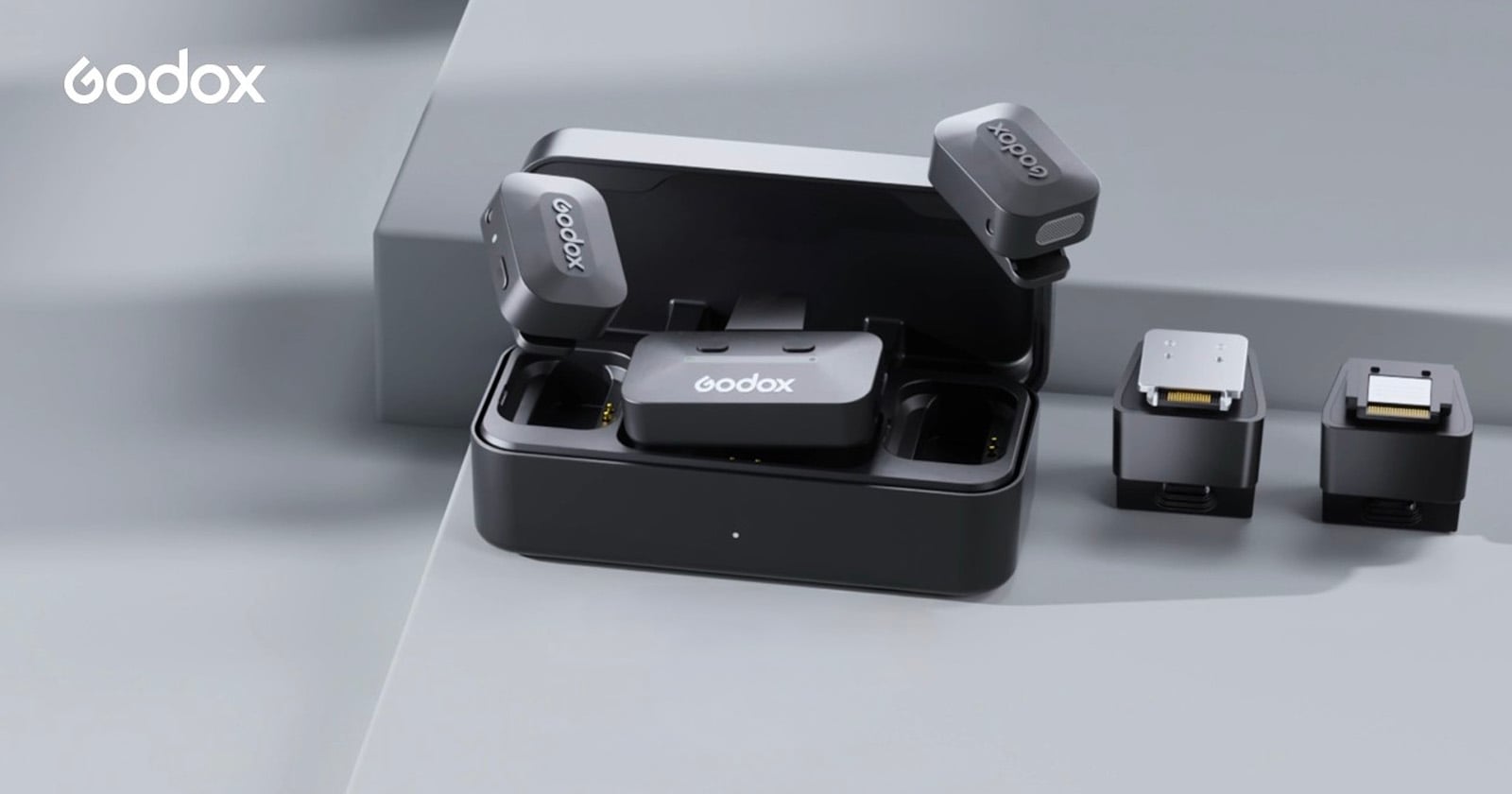 A Godox wireless microphone set is displayed on a gray surface, including a charging case, two microphones, and two receivers, with the Godox logo visible in the top left corner.