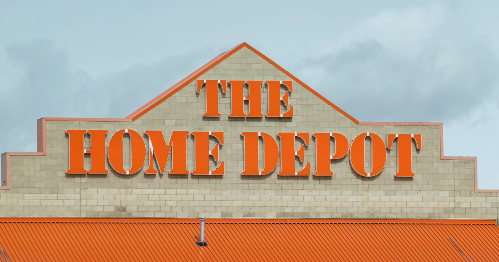 The Home Depot store exterior with large orange letters spelling The Home Depot on a beige brick wall and an orange roof below, set against a cloudy sky.