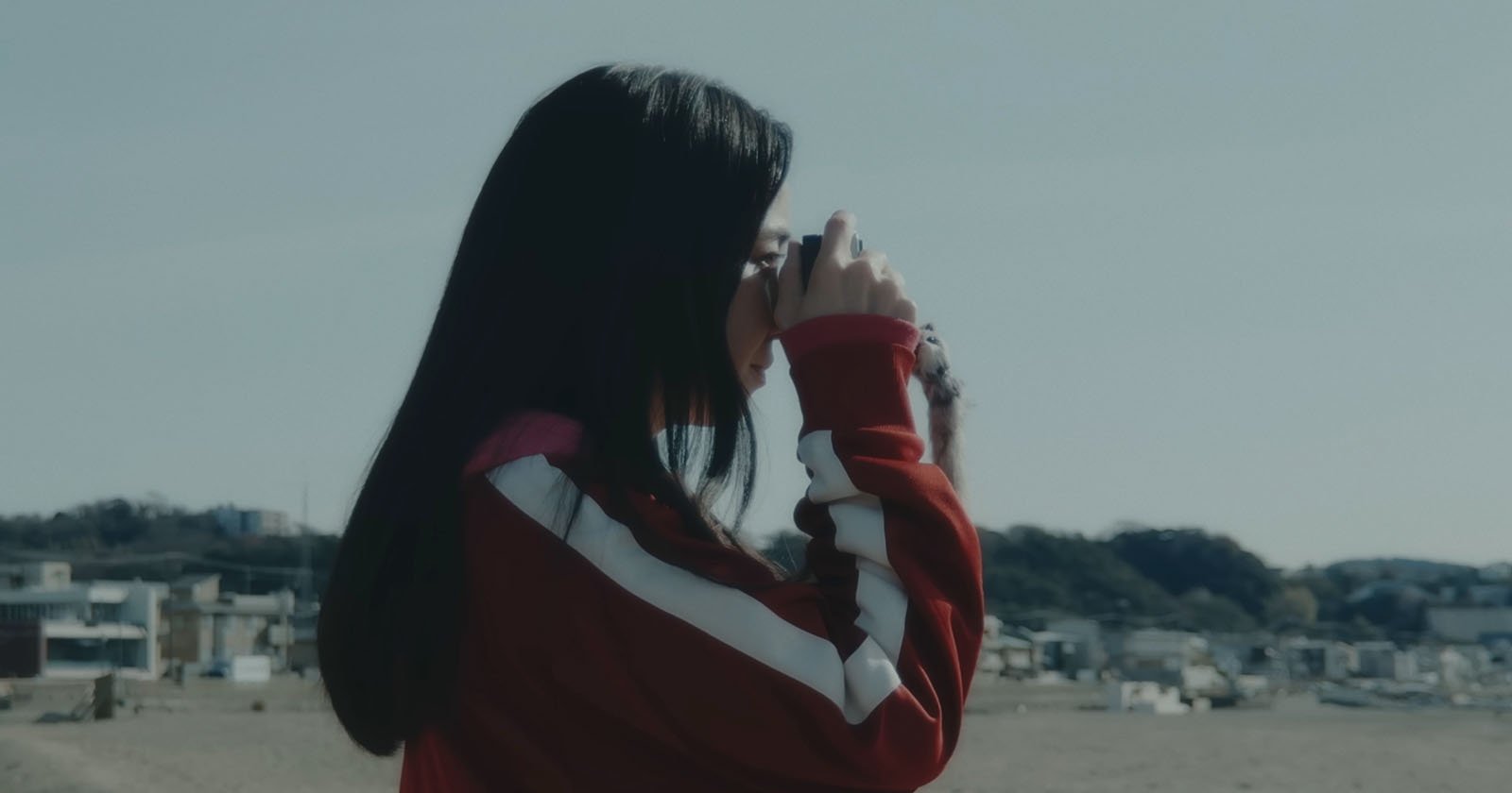 A person with long dark hair wearing a red and white jacket stands outdoors, holding a camera up to their face, with a blurred landscape and buildings in the background.