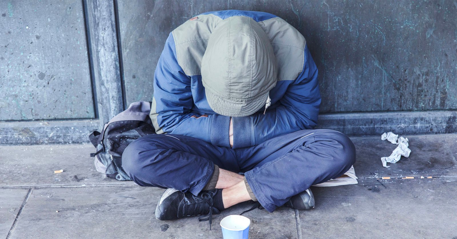 A person wearing a hooded jacket sits on the pavement with their head down and arms crossed over their knees. A backpack and a paper cup are nearby, and the background is a worn, gray wall.