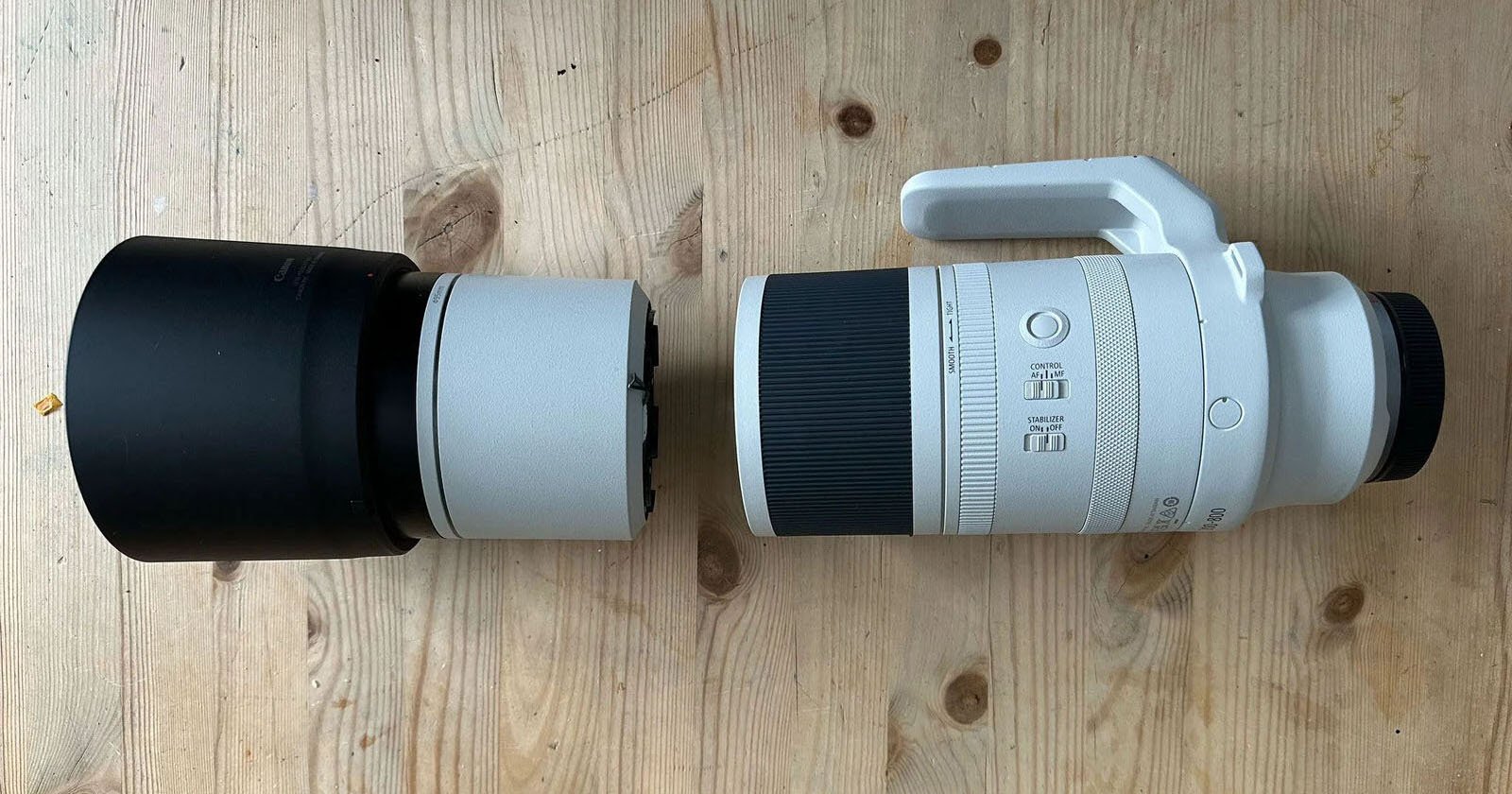 Two camera lenses on a wooden surface. The lens on the left is black and smaller, while the one on the right is larger, white, with a black grip and a tripod mount. Both are positioned horizontally.