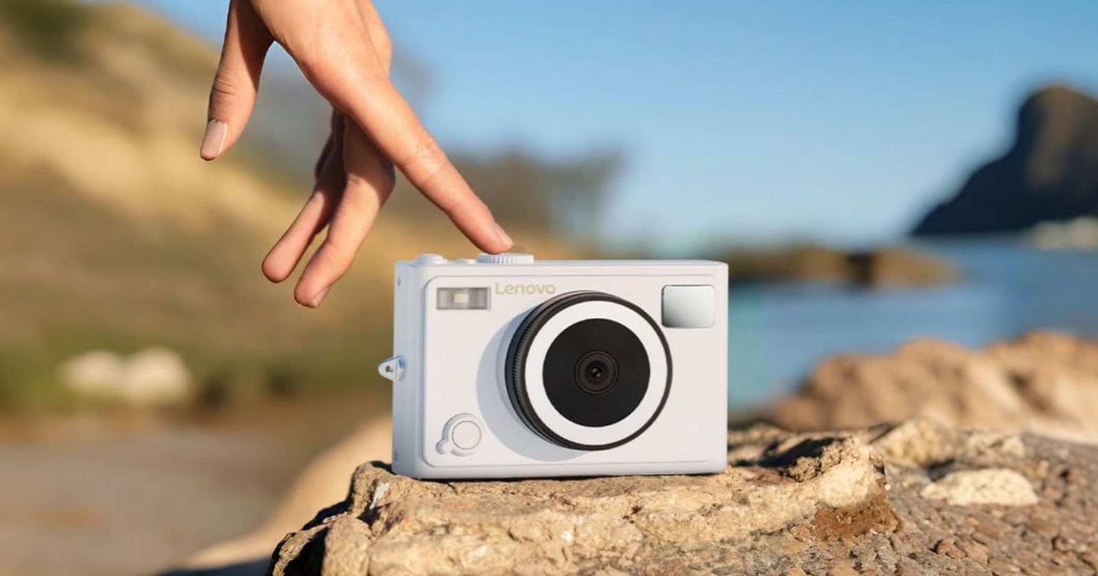 A hand gently touches a white Lenovo camera resting on a rock outdoors, with a blurred natural landscape and blue sky in the background.