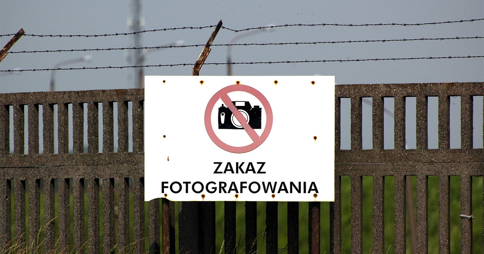 A weathered sign on a metal fence displays a crossed-out camera icon and the text Zakaz Fotografowania, indicating no photography allowed. The fence is topped with barbed wire, and greenery is visible in the background.