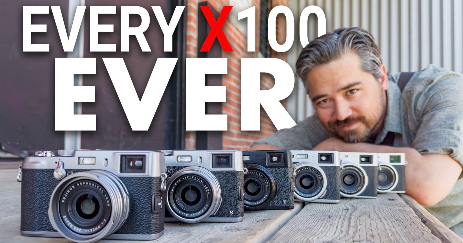 A person with grey hair and a beard is leaning on a wooden table, smiling at the camera. In front of them, there are six different Fujifilm X100 series cameras lined up. The text above reads EVERY X100 EVER. The background has an industrial look with brick and metal elements.