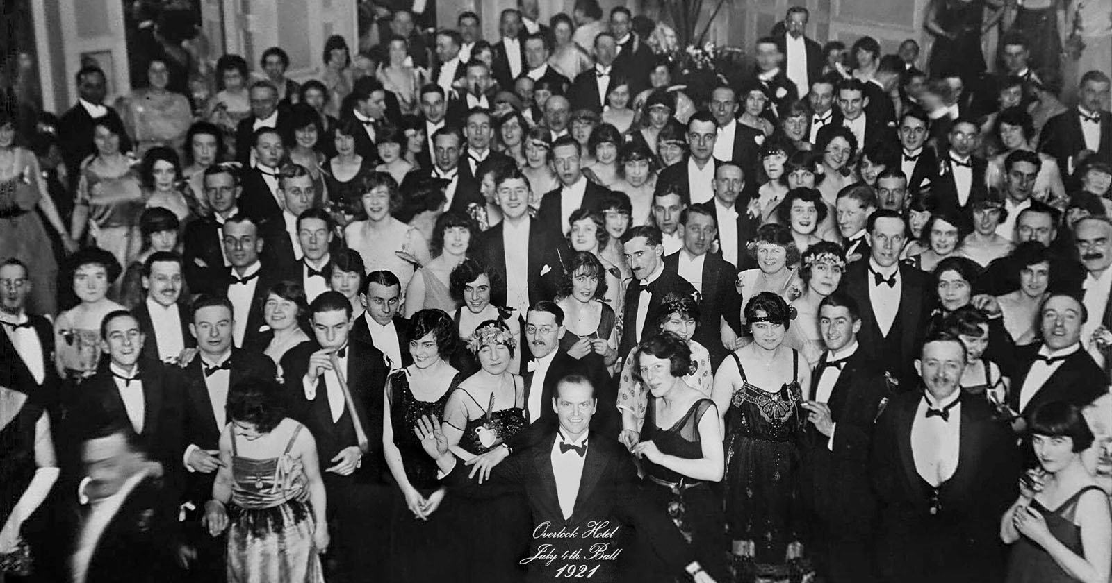 A black and white photo of a large group of people at a formal gathering in 1927. Many are in tuxedos and evening dresses, smiling and facing the camera. The setting suggests a grand ballroom atmosphere.