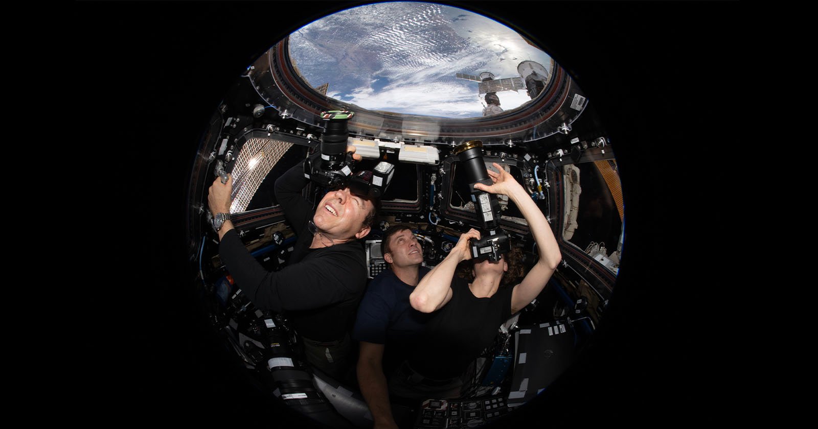 Three astronauts inside the International Space Station's cupola, capturing images of Earth using cameras. The window offers a striking view of the planet's surface. They appear focused, with one pointing and the others looking through their cameras.