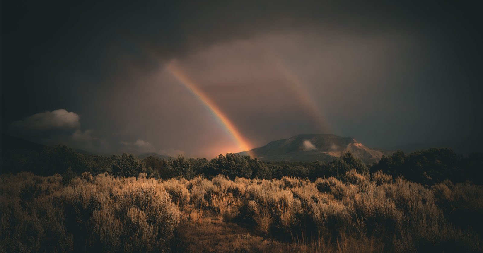 A dramatic landscape featuring a dark, stormy sky over a hilly terrain with a double rainbow arching over the scene. Grassy fields and scattered bushes dominate the foreground, and the distant hills are partially obscured by clouds.