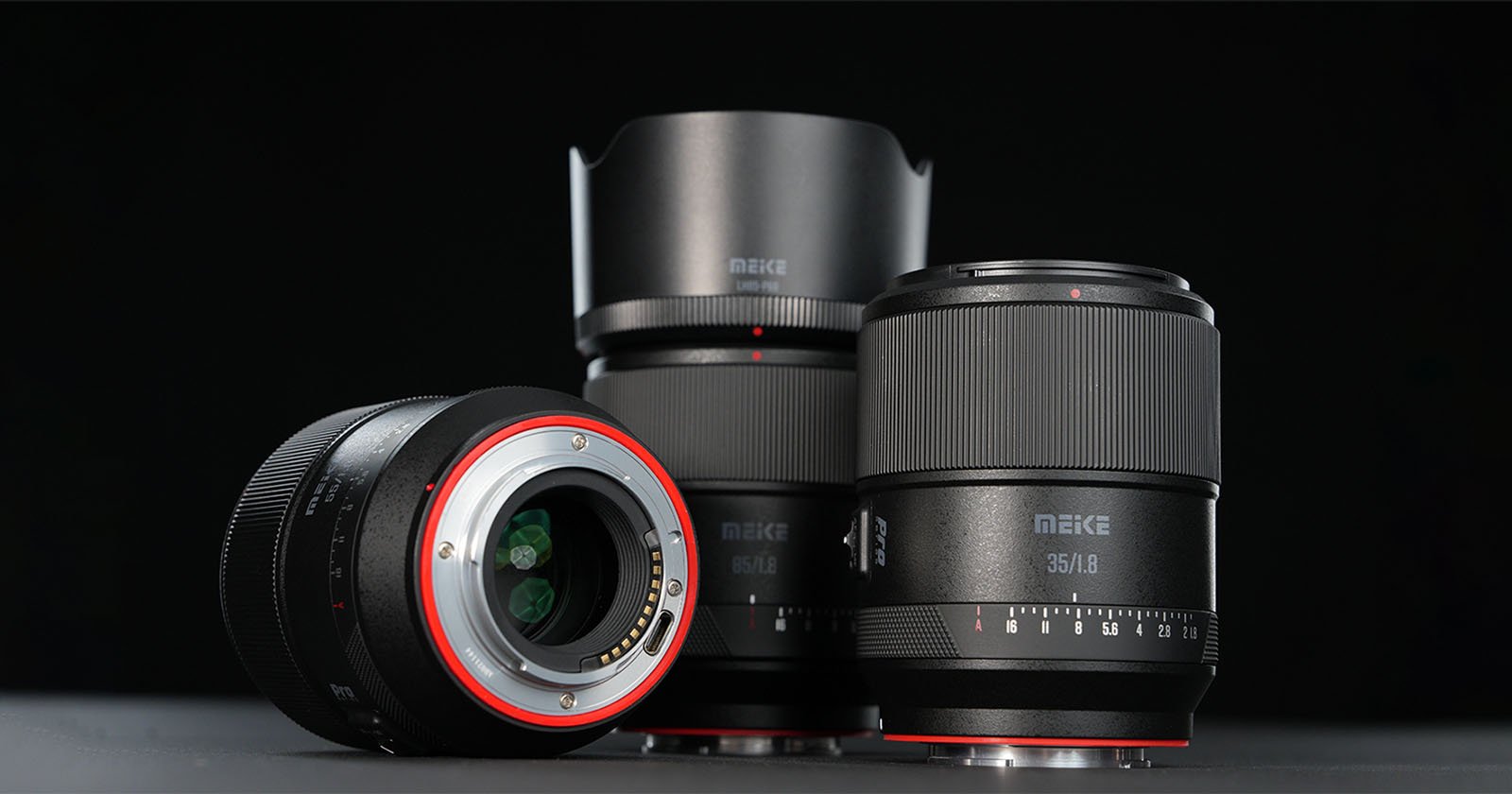 Three Meike camera lenses are displayed on a dark surface with a black background. One lens is lying down showing the mount, while the other two stand upright, highlighting their textured focus rings and branding.