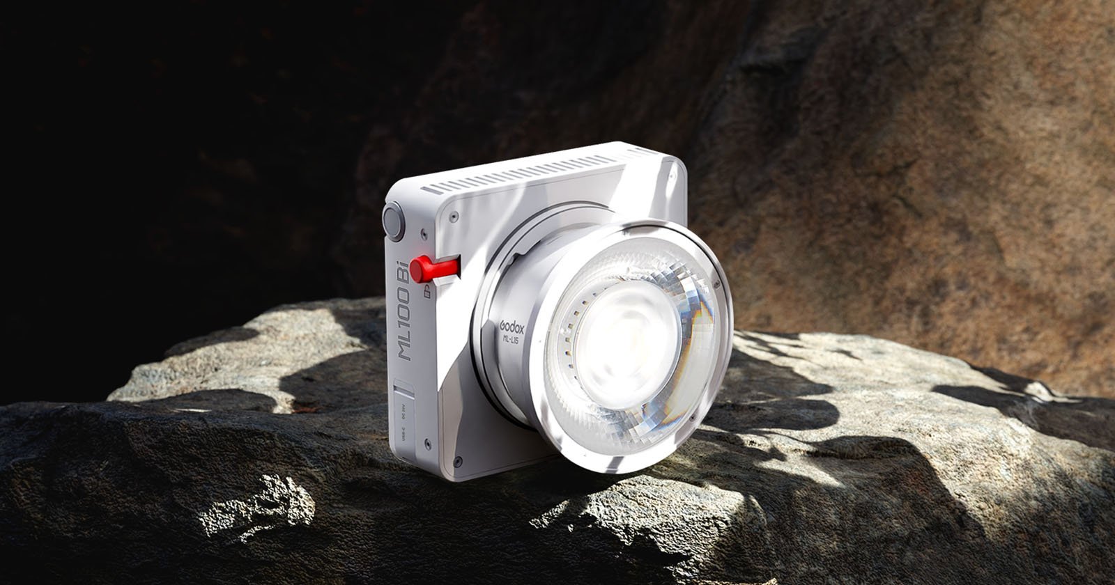 A modern white digital camera labeled ML100 with a large lens is positioned on a rocky surface, partially illuminated by sunlight. The background consists of uneven, natural rock formations.