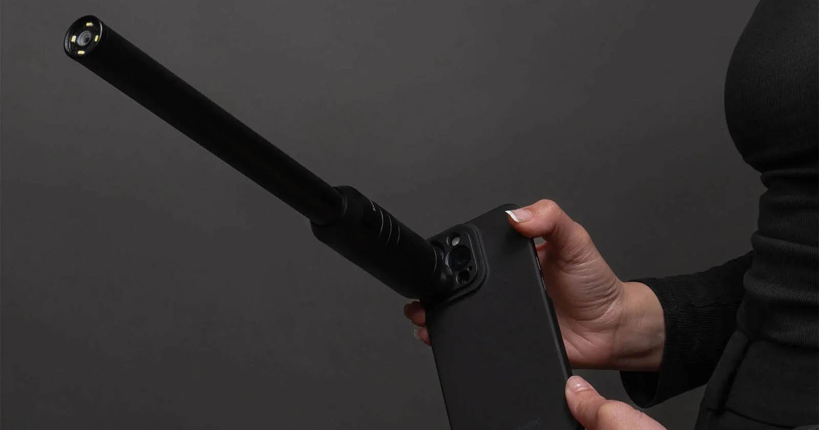 A person holds a black smartphone with an attachment that resembles a long camera lens. The background is dark, and the person's hands and part of their torso are visible.