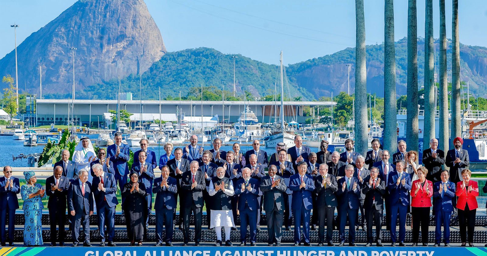 A large group of people stands on a platform, clapping, with a marina and a large mountain in the background. The stage banner reads Global Alliance Against Hunger and Poverty. The scene is sunny and clear.