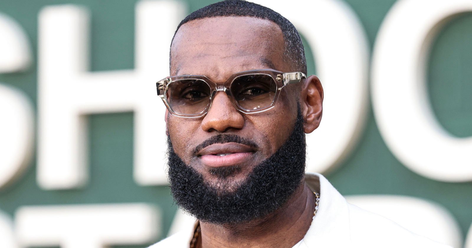 A man with a neatly groomed beard and mustache, wearing stylish sunglasses and a white shirt, stands in front of a green background with large, blurred white letters.