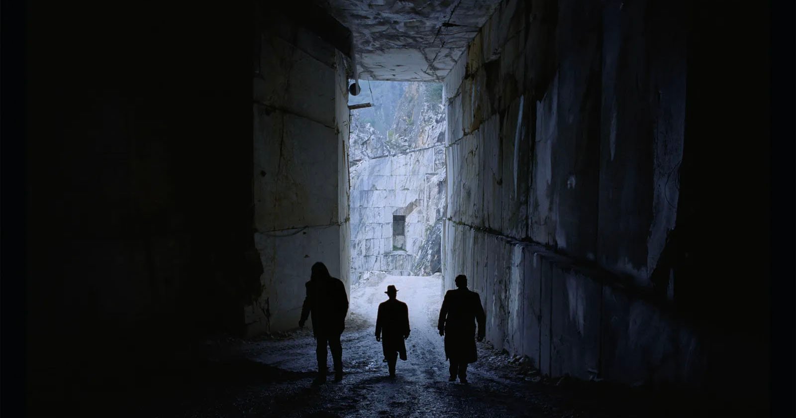 Three silhouetted figures walk through a dimly lit stone tunnel, with light visible at the far end. The central figure wears a distinctive hat, creating a mysterious atmosphere. Stone walls tower on both sides.