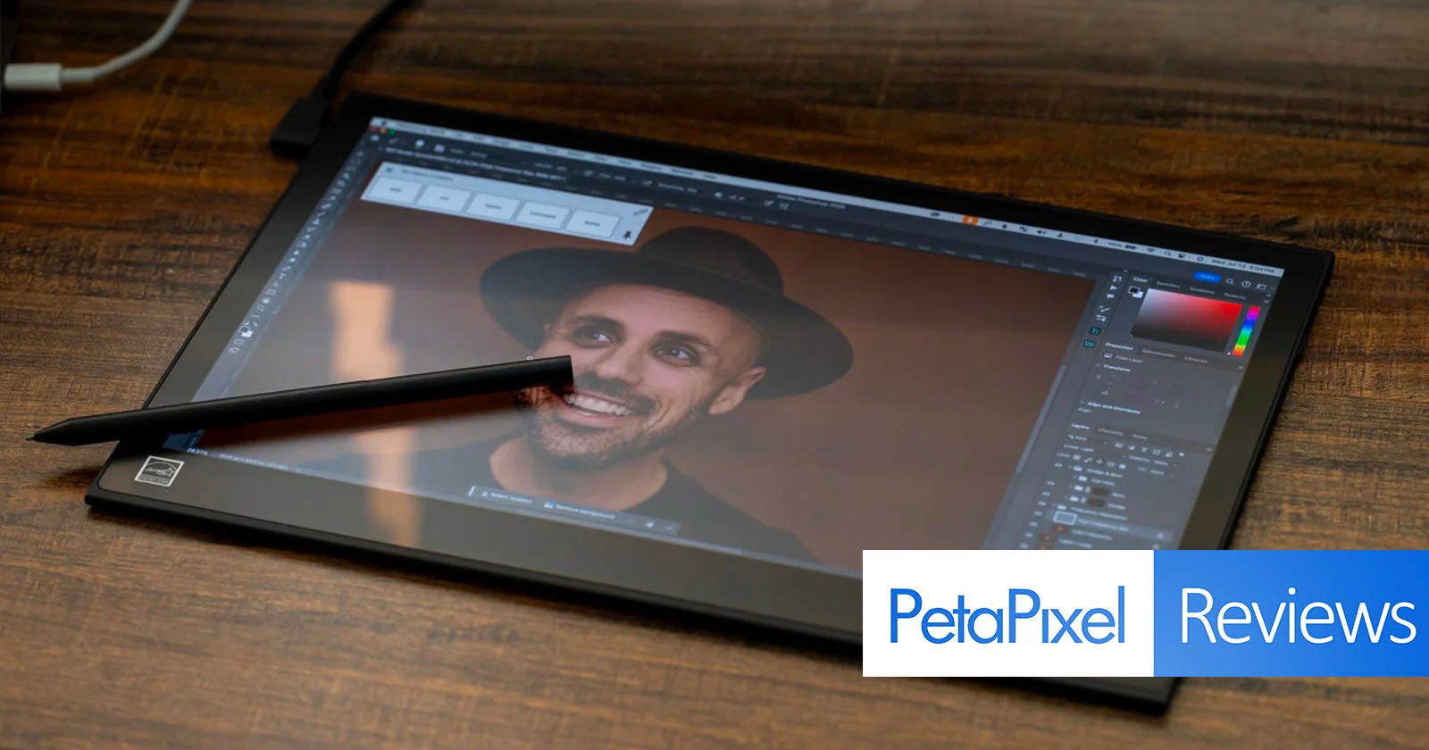 A digital pen rests on a tablet screen displaying a photo editing software with an image of a smiling person wearing a hat. The tablet is placed on a wooden surface. The bottom right corner features the PetaPixel Reviews logo.