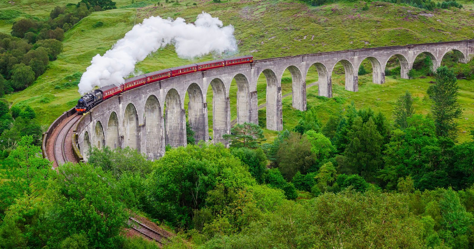 A steam train with red carriages crosses a tall, curved stone viaduct surrounded by lush green trees and rolling hills, with white smoke billowing from the engine.