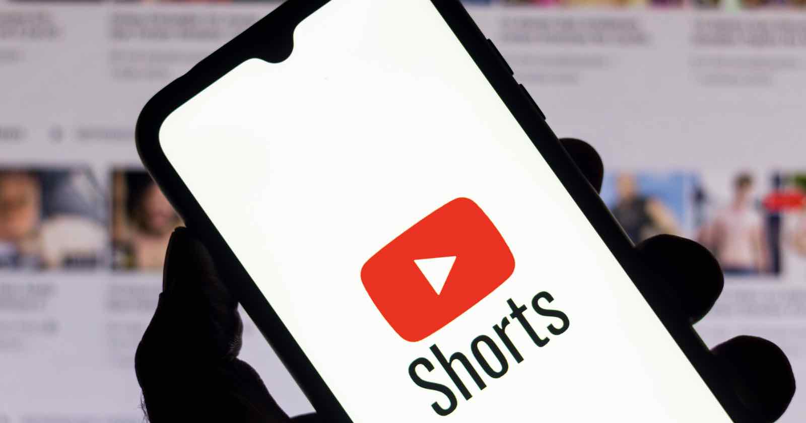 A hand holds a smartphone displaying the YouTube logo with the word Shorts below it. The background shows a blurred screen with thumbnails of multiple videos.