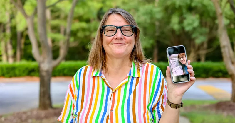 A person with shoulder-length hair and glasses is standing outdoors and smiling. They are holding up a smartphone displaying a photo of someone who appears to be the same person. They are wearing a colorful, striped shirt. The background includes trees and greenery.