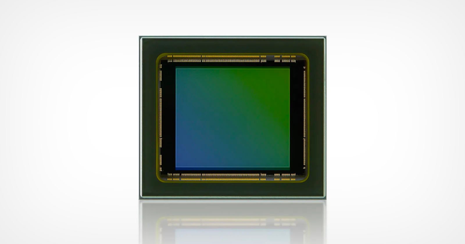 Square image sensor with a gradient from green to blue in the center, surrounded by a black border with gold contacts. The sensor is set against a light background, and its reflection is visible below.