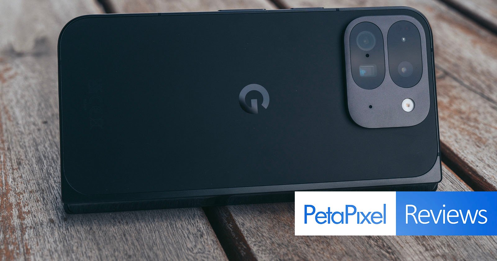 A smartphone with a dual-camera setup is placed on a wooden surface. The phone is black with a distinctive logo in the center. The bottom right corner has a PetaPixel Reviews logo.