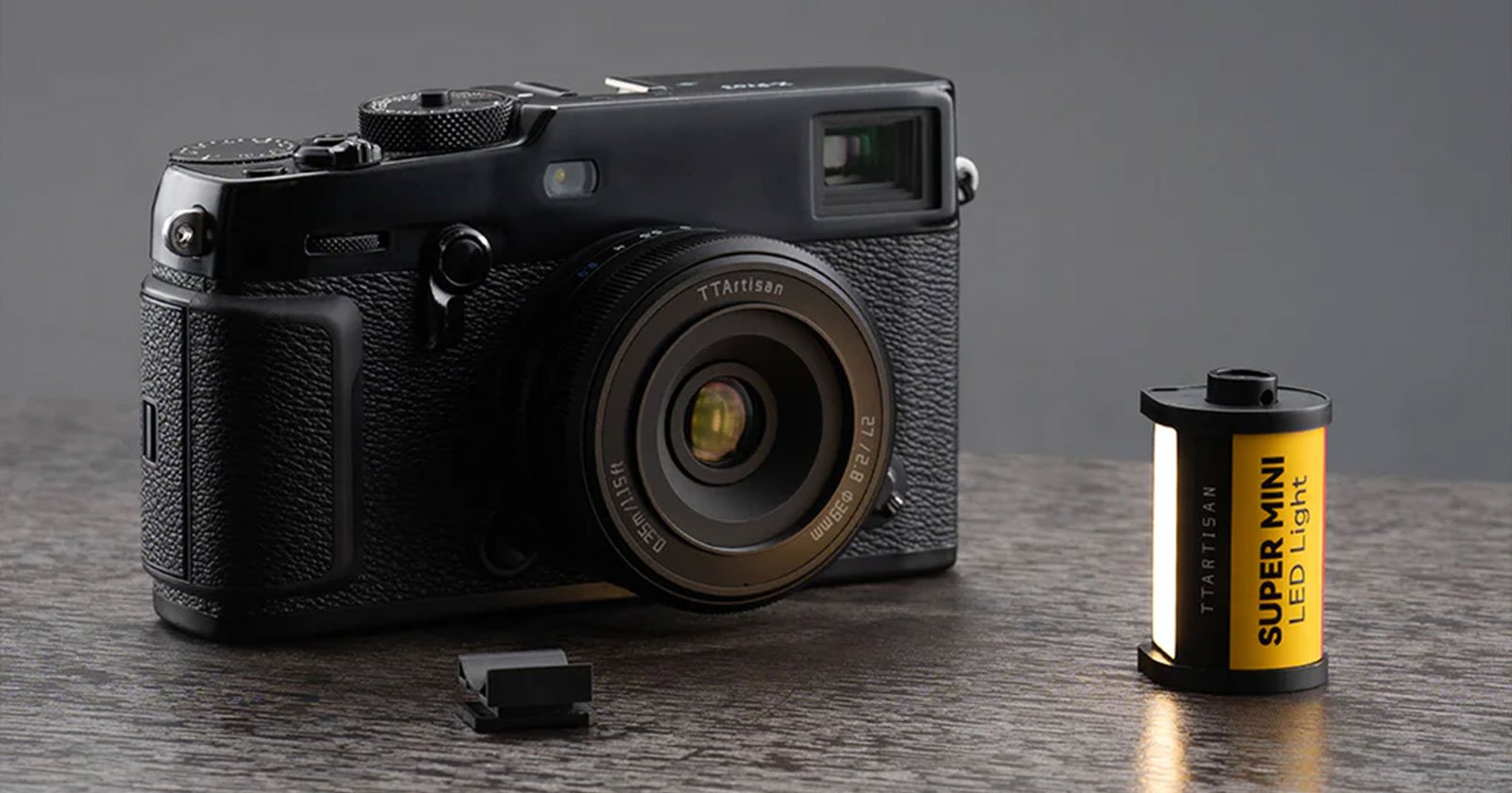 A black vintage-style camera sits on a wooden surface with a lens cap placed next to it. Beside the camera is a small roll of yellow film labeled SUPER-MINI LCD 100R with black and white text. The background is a plain, dark grey.