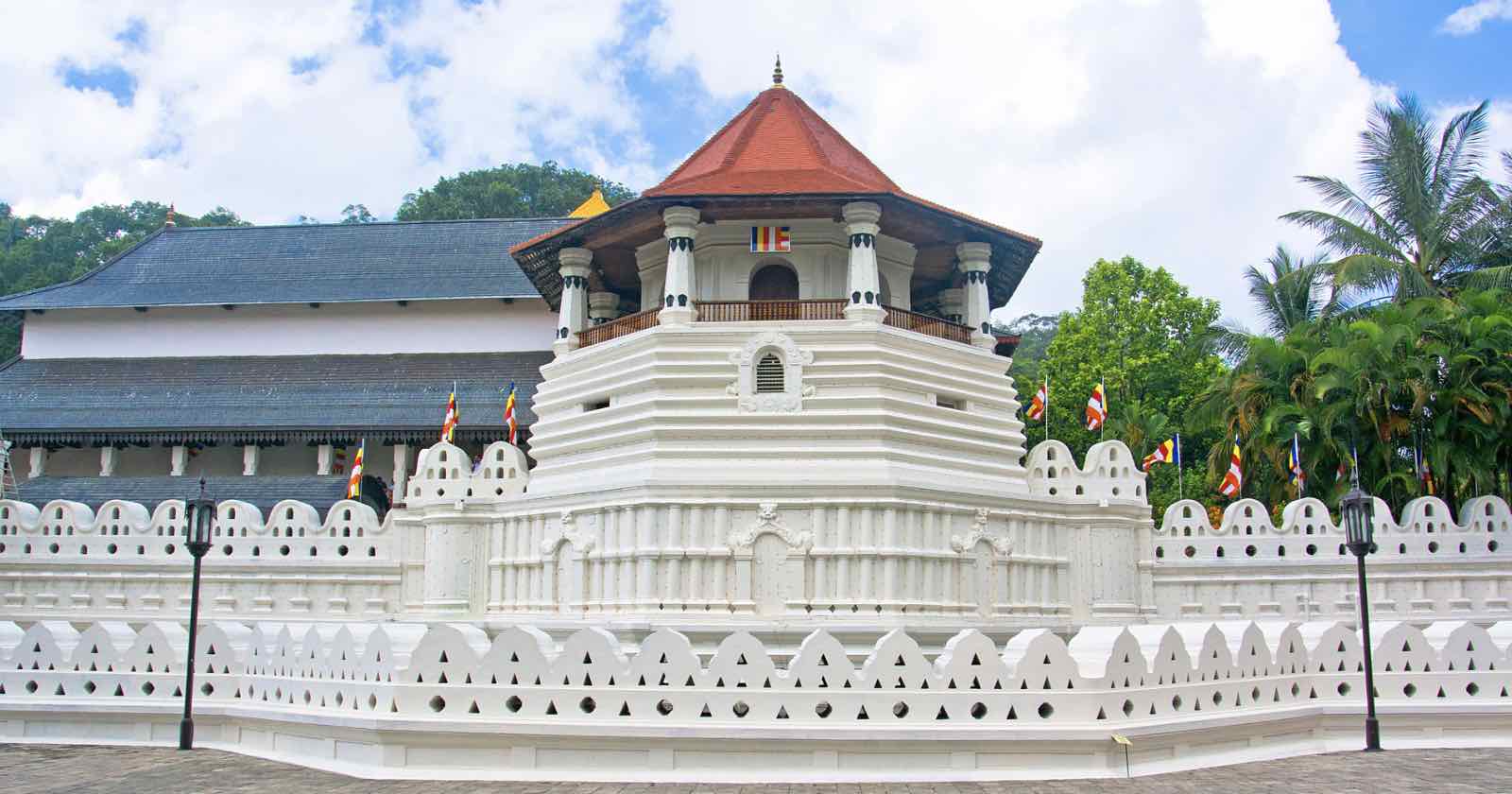 A white-walled temple with a red-tiled roof and small flags, surrounded by decorative walls and greenery, under a partly cloudy sky.