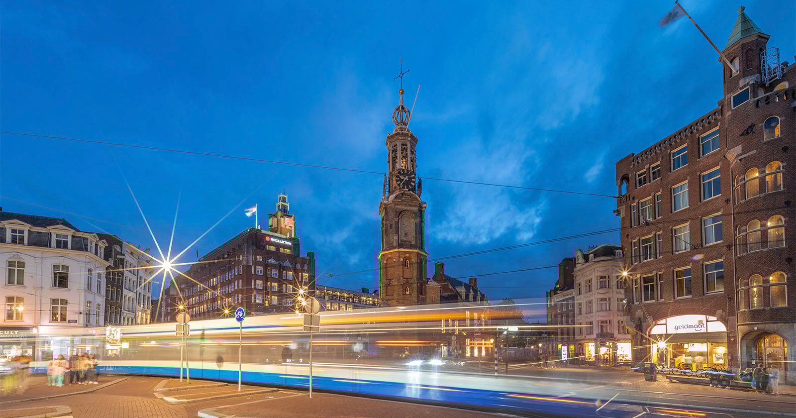 A cityscape at dusk shows blurred lights from a passing tram in front of historic buildings and a tall clock tower, with a deep blue sky in the background.