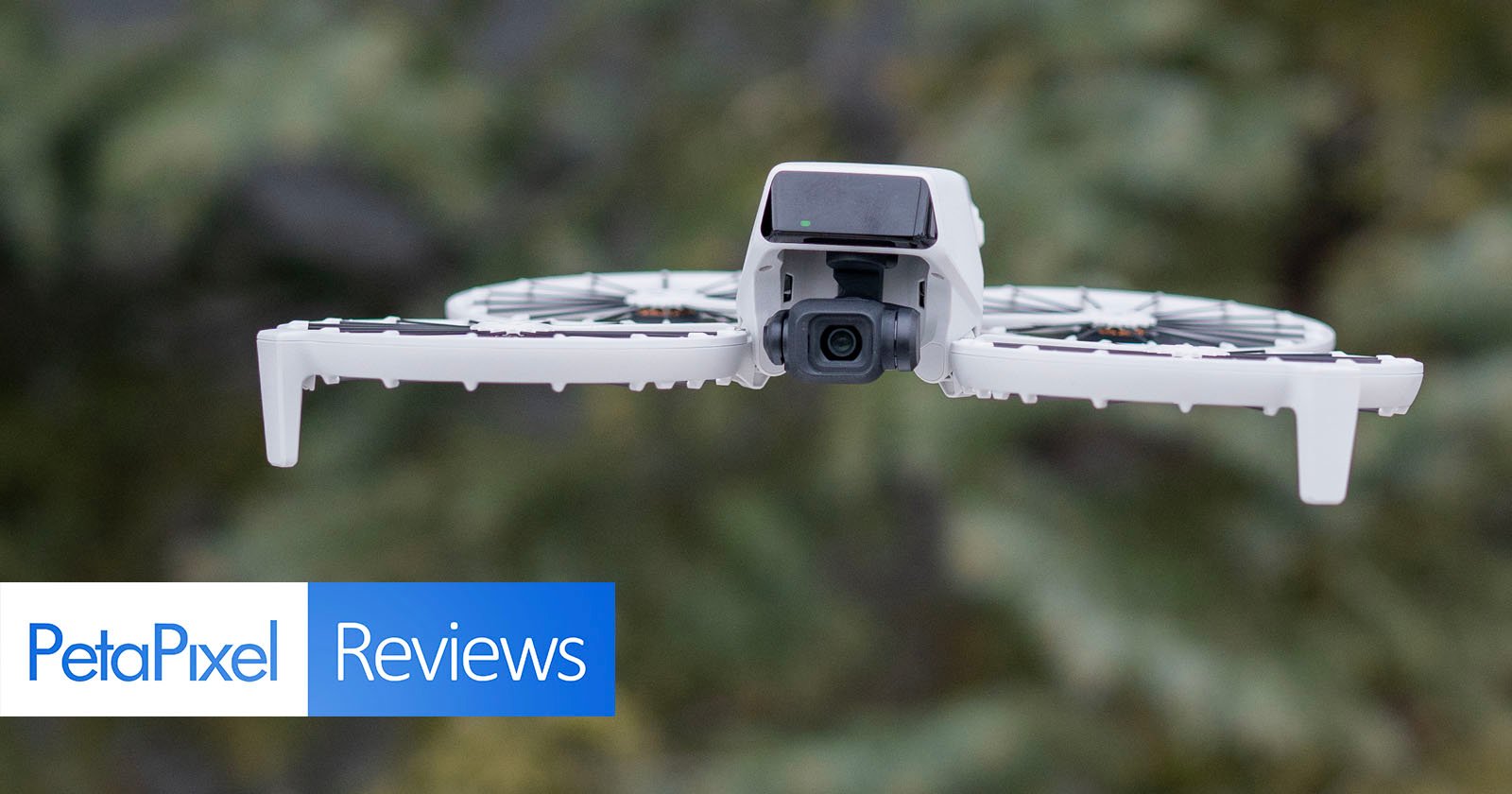 A drone with a camera flying outdoors against a blurred green background. The image has text reading PetaPixel Reviews in the bottom left corner.
