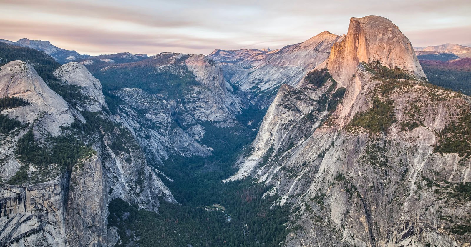 A dramatic view of Yosemite Valley with steep granite cliffs, dense forest below, and the iconic Half Dome rising prominently under a soft, pastel sky at sunset.