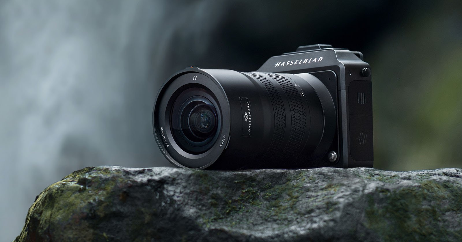 A black Hasselblad camera with a large lens rests on a moss-covered rock in an outdoor setting. The background is blurred, emphasizing the camera's details, including the textured grip and labels on the lens and body.