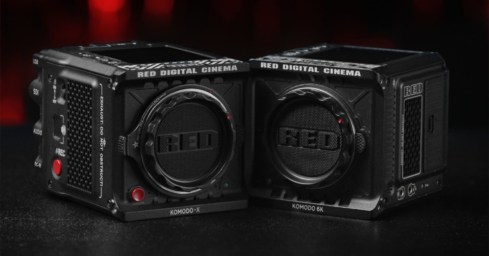 Two black RED Digital Cinema Komodo-X and Komodo 6K cameras sit side by side on a dark surface with a red and black blurred background. The lenses prominently display the RED logo.