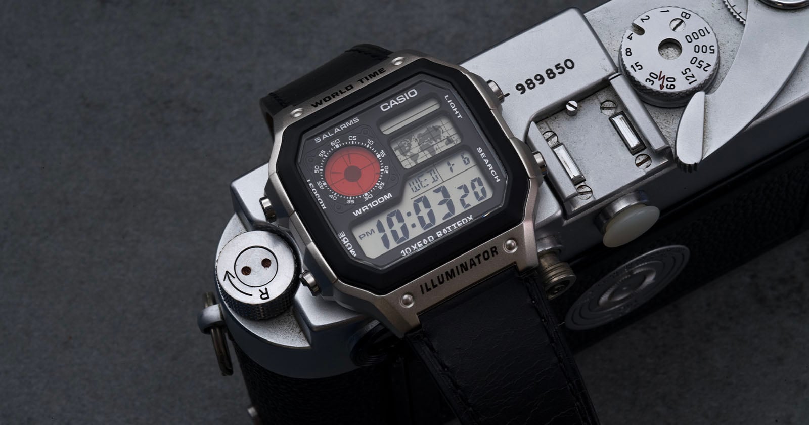 A Casio digital wristwatch with a black strap and multiple displays rests on top of a vintage silver camera on a dark surface. The watch face shows the time as 10:03:20 and features world time and illuminator functions.