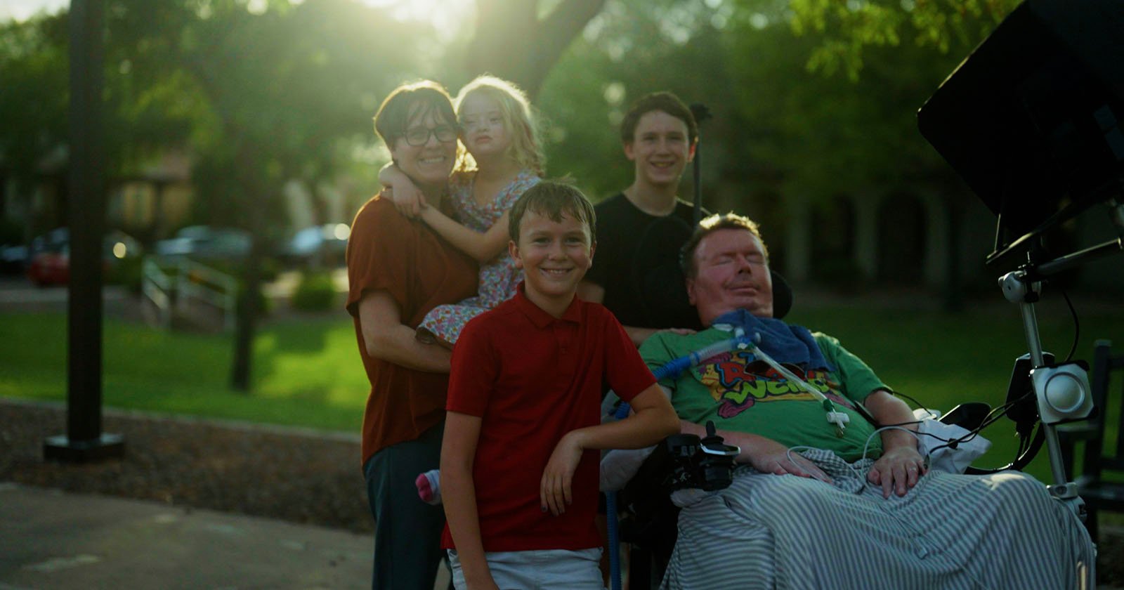 A group of five people, including a child, stand and smile outdoors near a man in a wheelchair equipped with medical equipment. Sunlight filters through the trees in the background, creating a warm and happy atmosphere.