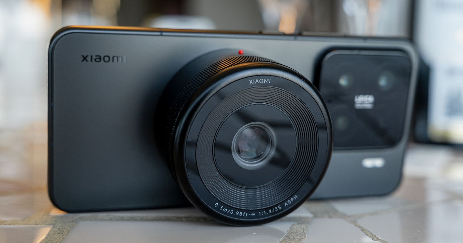 A smartphone with a large, detachable camera lens attachment displayed on a table. The lens covers part of the phone's back, giving it a DSLR-like appearance. The phone is branded with Xiaomi.