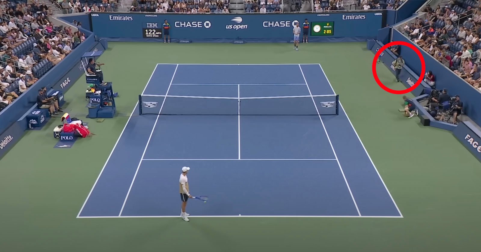 A wide view of a tennis match at the US Open with one player on the court. A person in the stands, circled in red, is leaning over the barrier. Spectators fill the stands, and the scoreboard is visible.