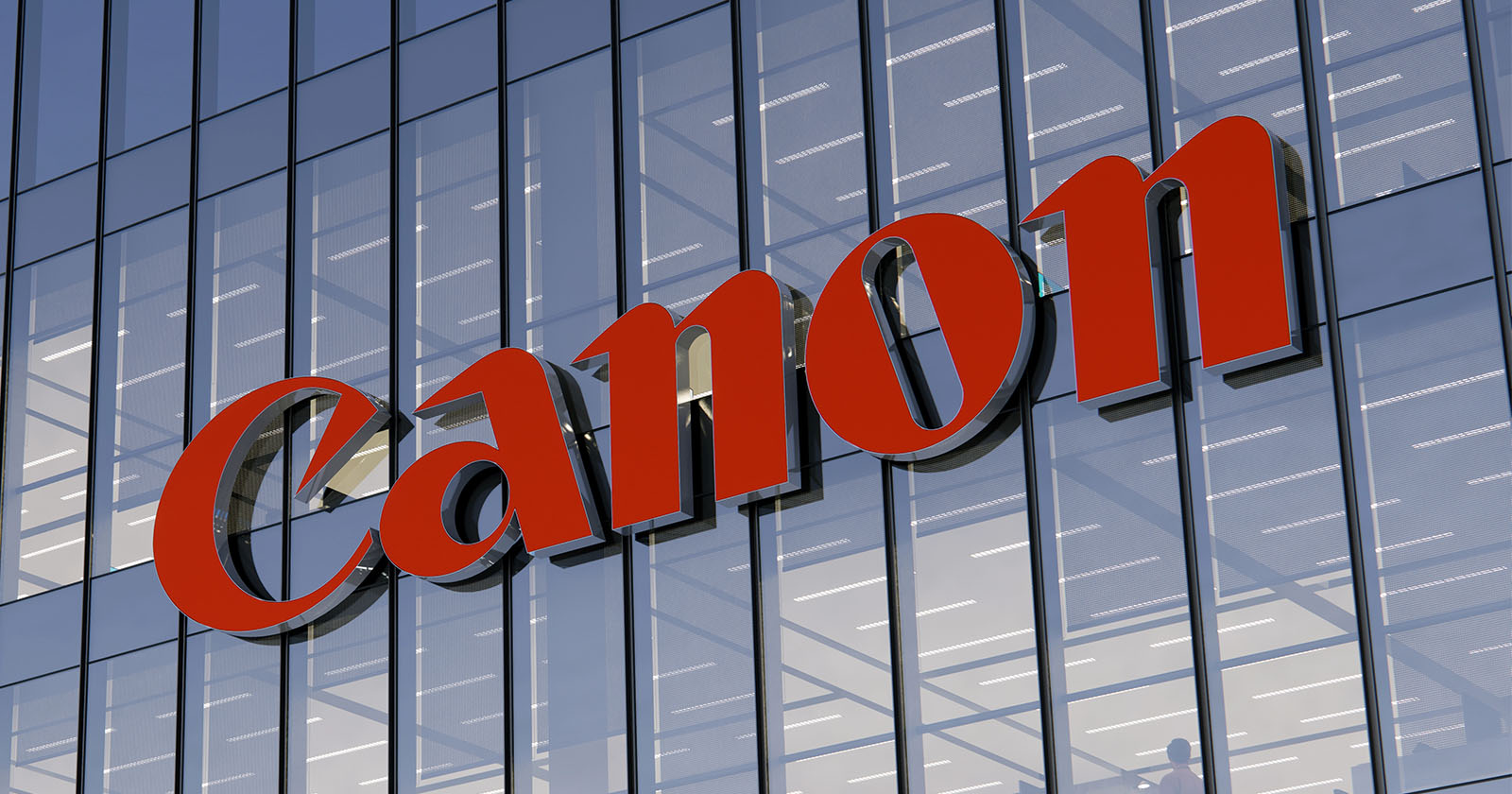 The image shows a glass building facade with a large red Canon logo prominently displayed. The reflective windows capture the sky and surrounding lights.
