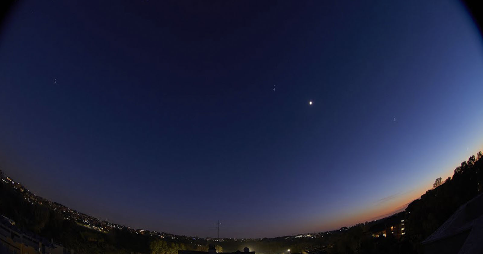 A twilight sky with a bright crescent moon and several stars visible. The horizon glows with the fading light of the setting sun. Below, a landscape of distant city lights and darkened silhouettes of trees is visible.