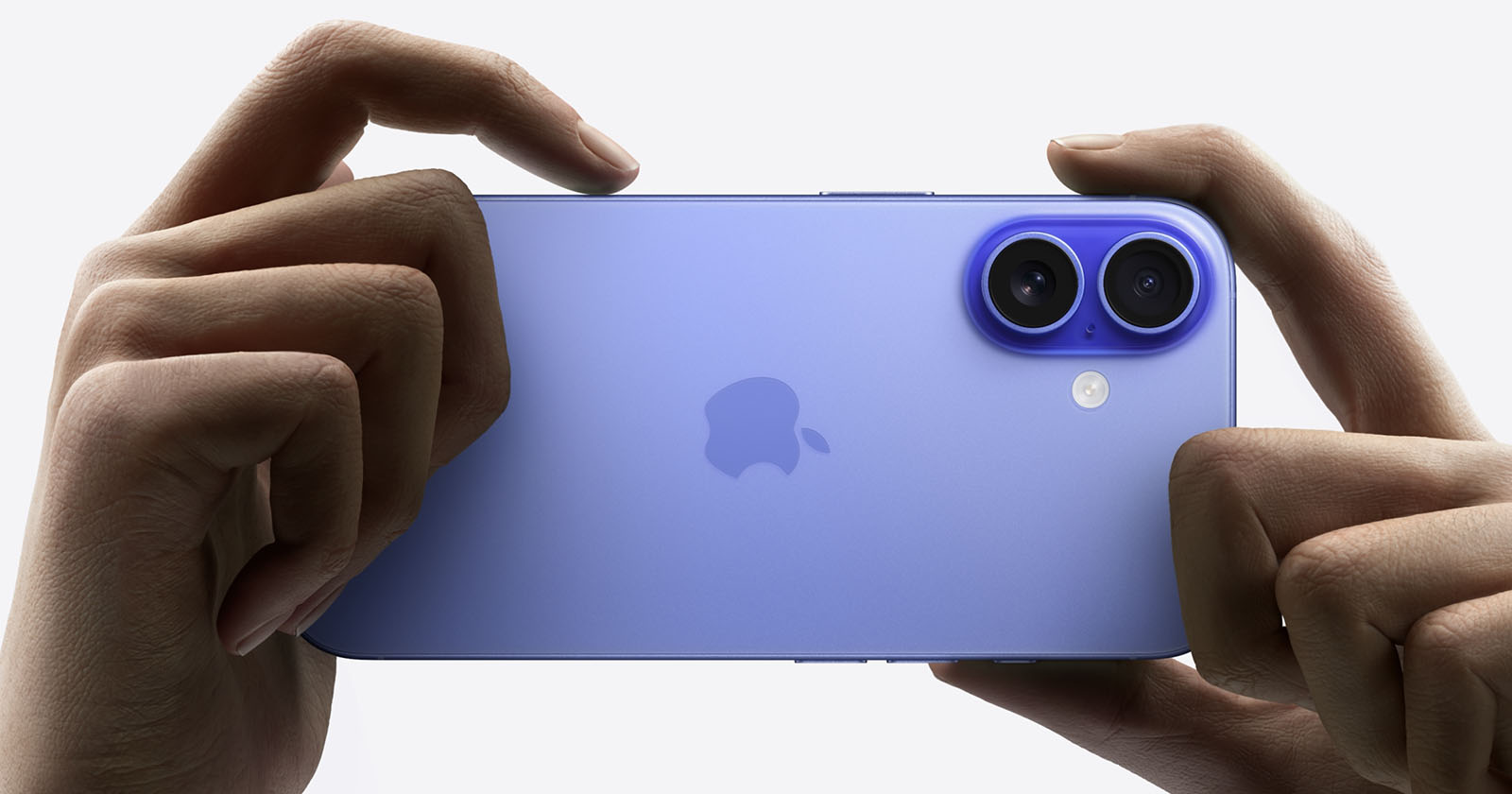 A pair of hands holding a purple iPhone horizontally. The phone has dual rear cameras and an Apple logo on the back. The background is white, emphasizing the phone's color and design.