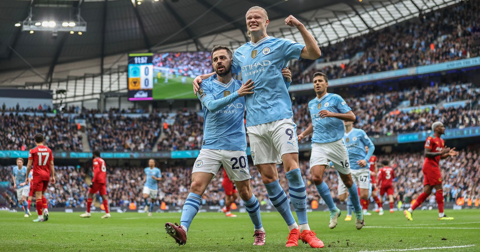 Manchester City footballer Erling Haaland celebrates after scoring a goal against Liverpool during a match at the Etihad Stadium. Teammates surround him as fans cheer from the stands, while Liverpool players are seen in the background.